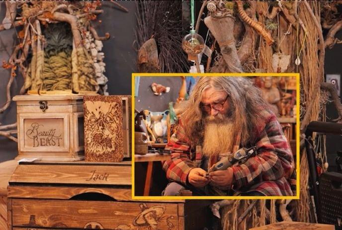 A bearded man in a plaid shirt uses a handheld tool, surrounded by wooden sculptures and carvings.