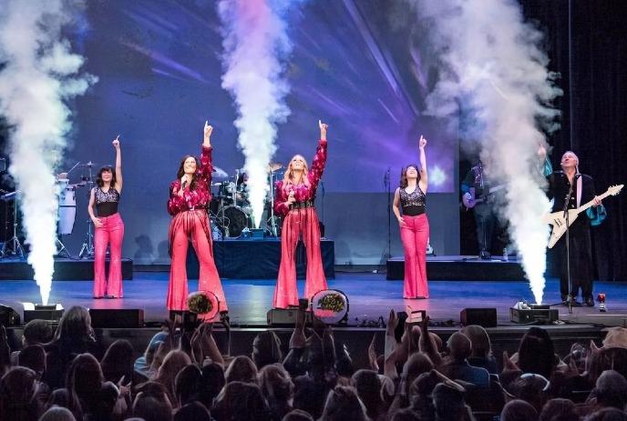 Four women in vibrant pink outfits and a guitarist to their left perform energetically on stage, pointing upward.
