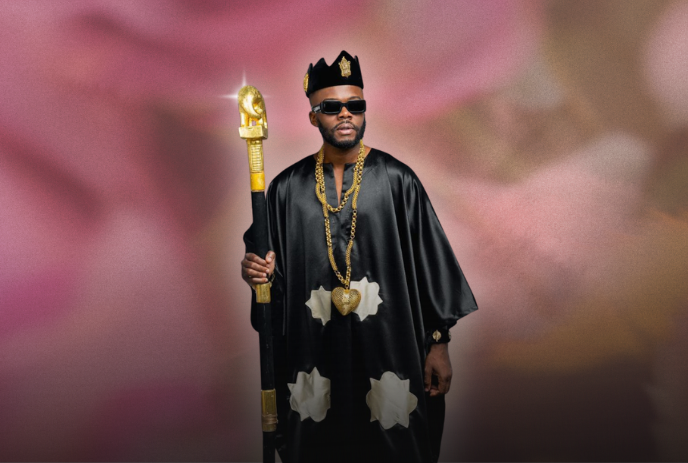 DJ Kerozen wearing sunglasses and holding a staff in front of a hazy pink background.