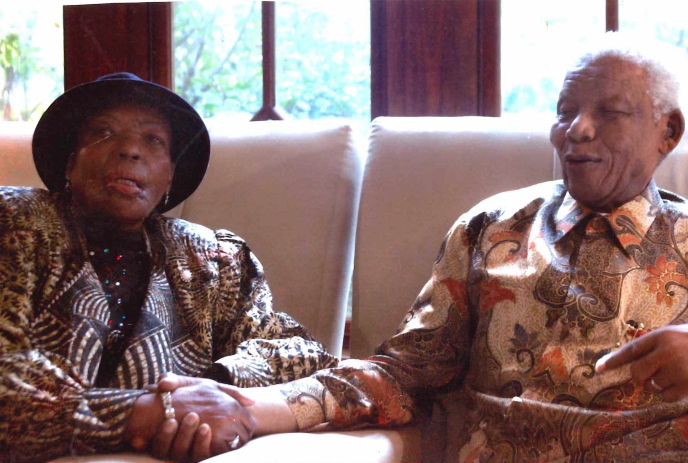Two older people sit close on sofa holding hands warmly lit room with large windows and gentle calm.