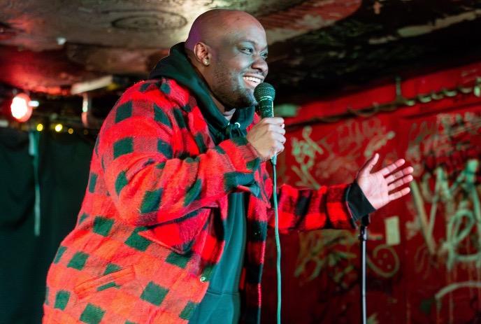 Gee Gethiga performing stand-up comedy on stage, smiling while holding a microphone in a red and green plaid jacket.