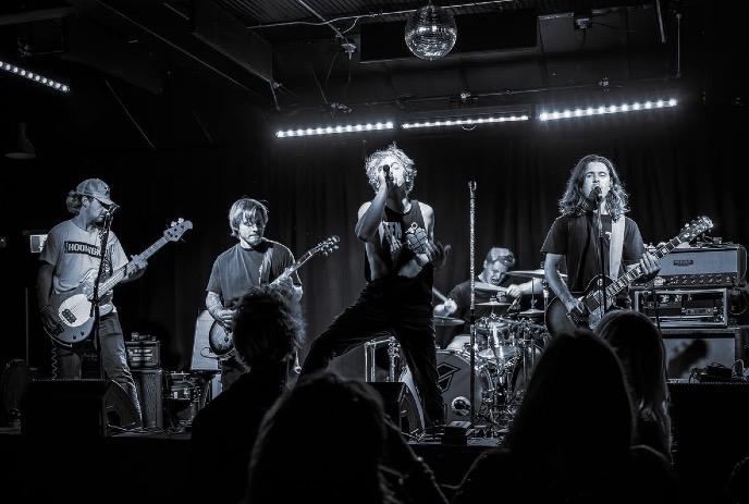 Five band members performing on the stage in a black and white frame.