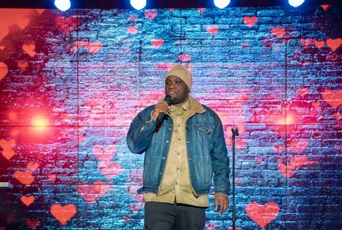 Performer on stage in denim jacket holds mic, hearts on brick wall screen glow under bright stage lights.