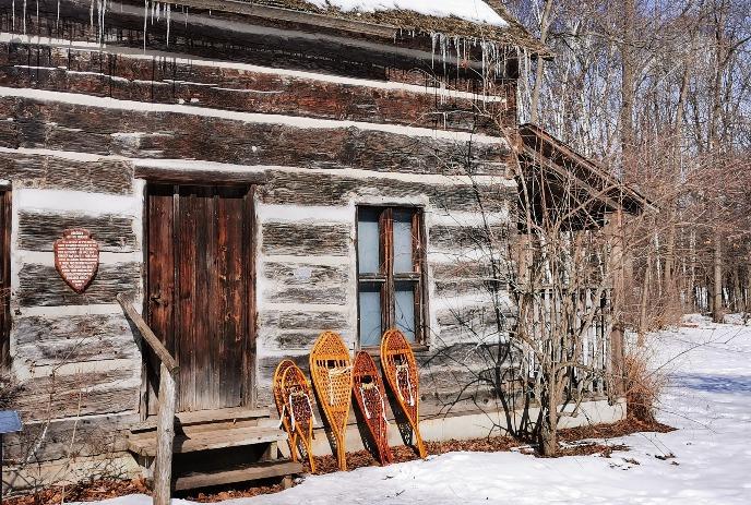 A weathered log cabin with icicles, set in snowy woods. Four rustic snowshoes lean against steps.