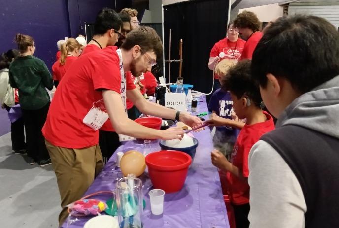 Kids explore a science table with dry ice fog as volunteers in red shirts guide hands?on experiments.
