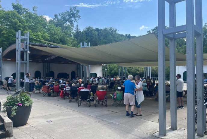 A large crowd gathers under a canopy in folding chairs at an outdoor amphitheater, surrounded by trees and a couple dancing.