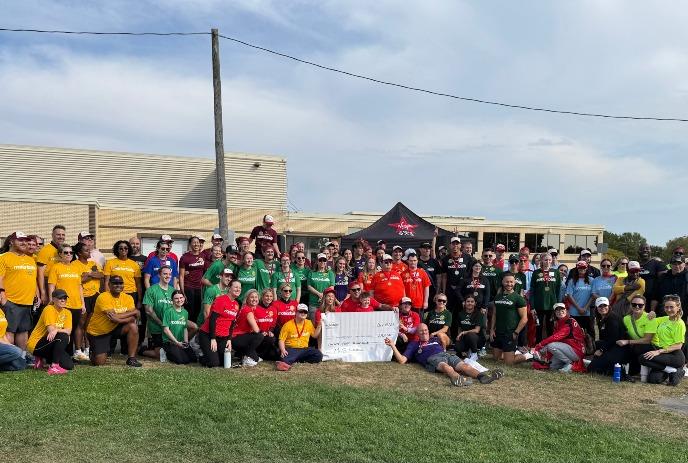 Large group in colored team shirts outdoors holding big sign with canopy tent and beige building behind.