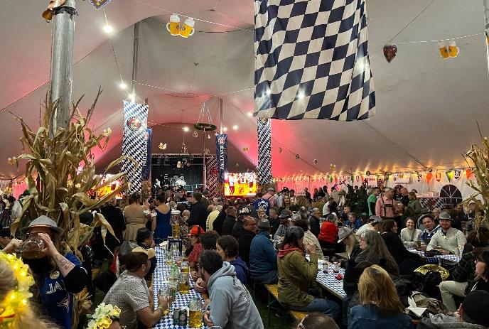 Crowded festive tent with long tables, bright decor and lively stage lighting during a beer celebration.