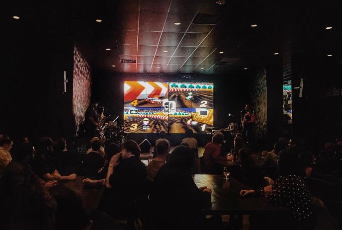 People playing Mario Kart in a dark room with a band on stage.