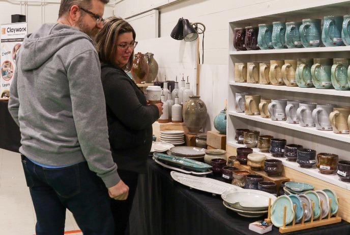 Two people browsing handmade ceramic pottery at a craft fair; shelves filled with colorful mugs, bowls, and vases.