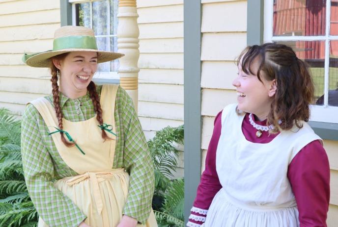 Two women in historical costumes stand outside a wooden house with ferns.