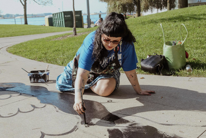 Natalie King painting on paved walkway outdoors with supplies nearby beside grassy area.