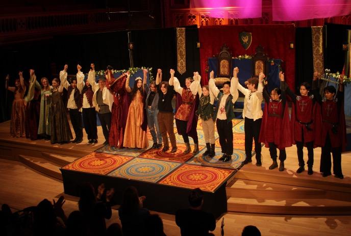 Actors in medieval costumes bow on ornate stage with thrones, banners, and warm lighting as crowd applauds.