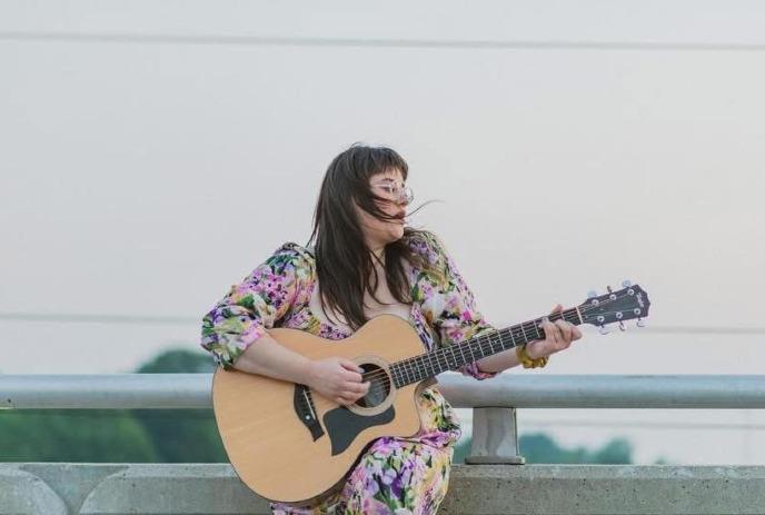 Artist sits on concrete ledge playing guitar as wind lifts hair against soft outdoor sky backdrop.