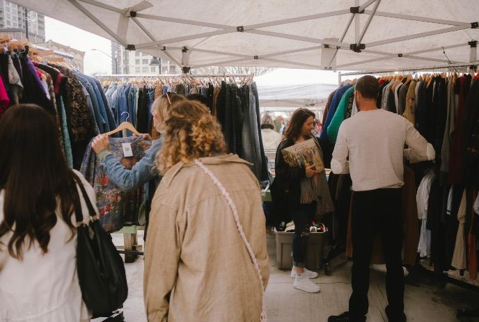 People browsing clothing racks at outdoor market stall under canopy.