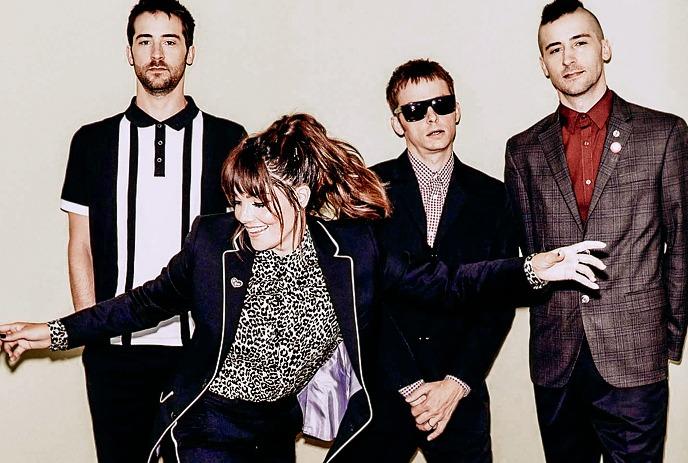 Four people pose against a plain backdrop, with one person in front extending both arms outward.