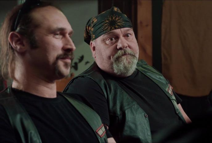 Two men in black leather vests sit together indoors, one wearing a patterned bandana.