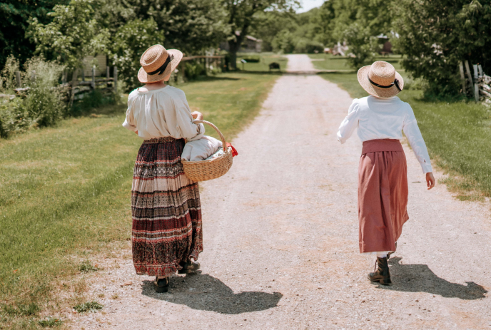 Two people in vintage clothing walk down a rural path, one carrying a basket.