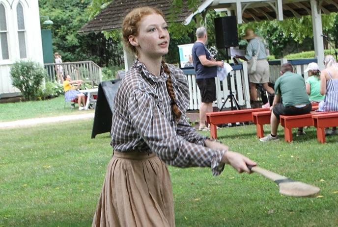 A young woman in 19th-century attire plays a lawn game at the Fanshawe Pioneer Village, with visitors gathered nearby.