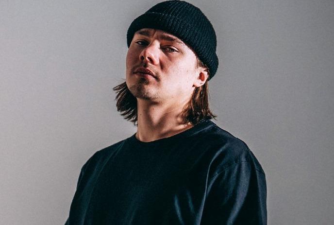 Izzy Vadim wearing a black hat and shirt, looking at the camera in front of a grey background.