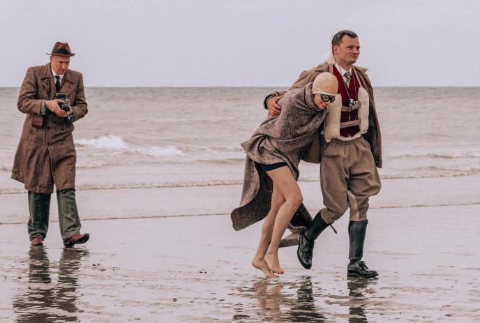 A young woman and a man walk along in the water with a photographer walking close behind them.