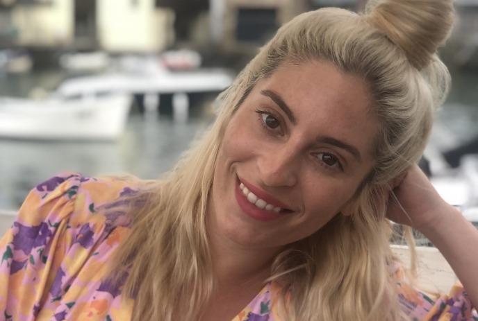 Jackie Pirico with light hair in a floral top, seated by a marina, leaning on one arm near boats.