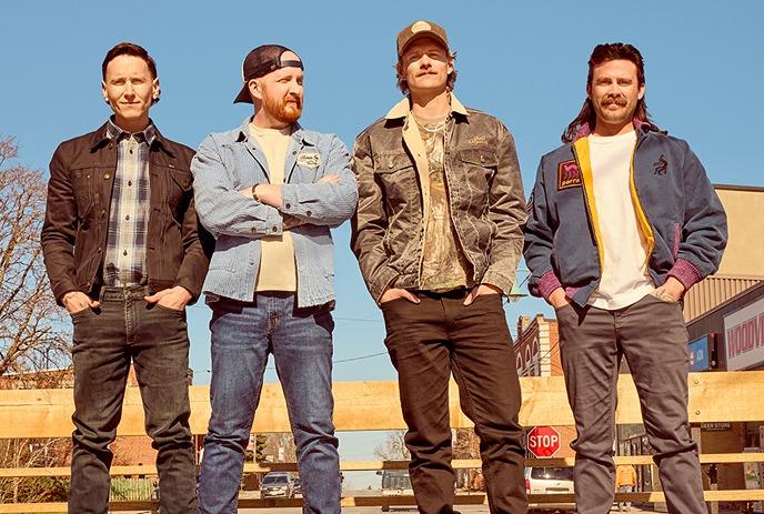 Members of The James Barker Band standing on a bench outdoors, wearing fall jackets