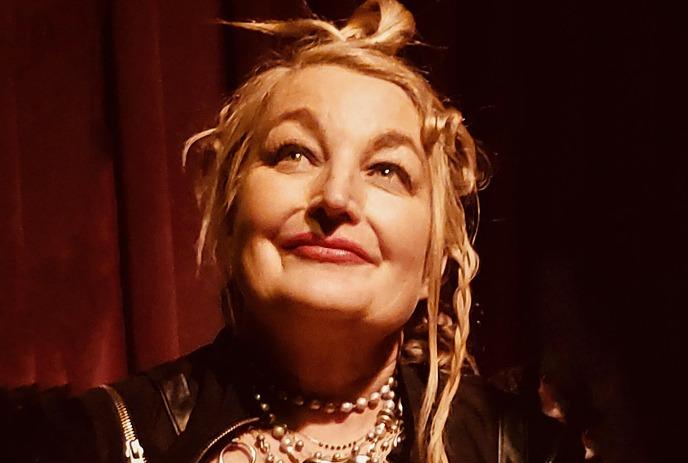 Jane Siberry with a radiant smile and blonde hair styled in an updo, wearing layered necklaces and a black outfit.