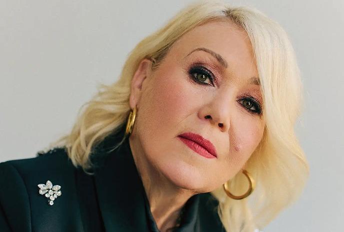 Portrait shot of Jann Arden with short blond hair in a black embroidered jacket, looking  forward.