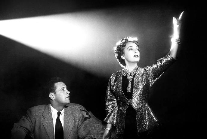 A diva is onstage in the spotlight while a man looks on.