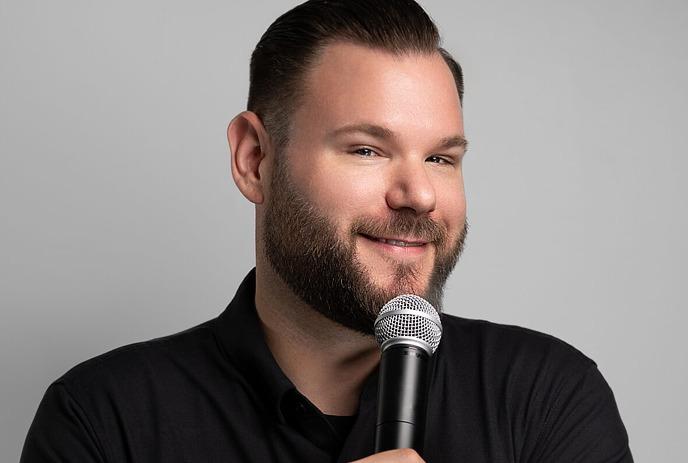 Jeff Leeson smiling in front of a grey wall with a microphone in his hand.