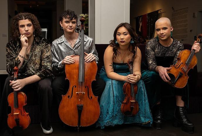 Four diverse musicians in sparkling attire sit with violins, viola, and cello in an art-filled room.