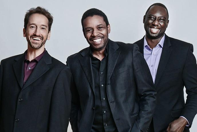 Three artists smile before a pale backdrop, wearing dark jackets and bright shirts.