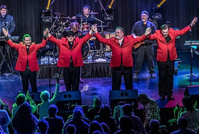 A band in red jackets holding hands up on a stage, cheered by a crowd.