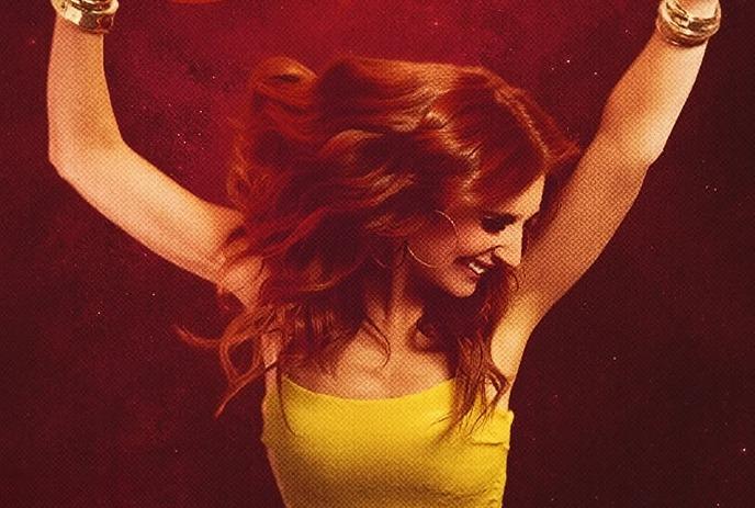 Artist with long wavy hair in yellow top arms raised against deep red backdrop suggesting lively motion.