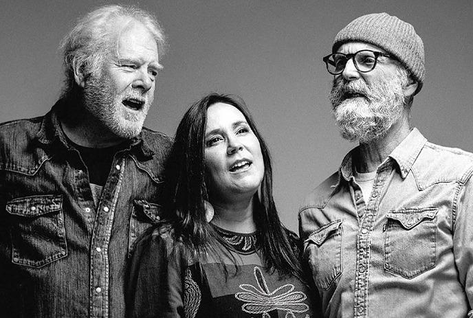 Three people standing together in a black-and-white portrait, appearing to sing or speak, dressed in casual clothing.