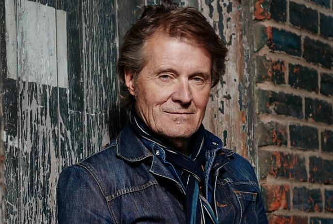 Jim Cuddy wearing a denim jacket posing for a picture under a bricked wall.