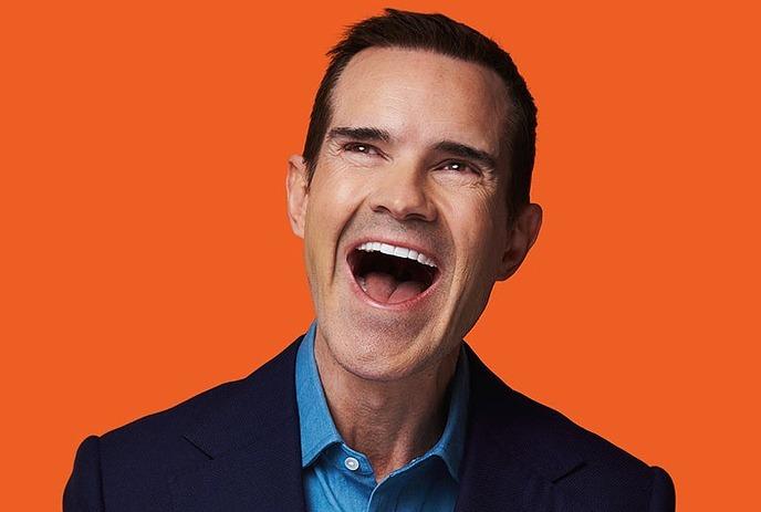 Jimmy Carr wearing a black coat and blue shit, smiling, in front of an orange maple leaf background.