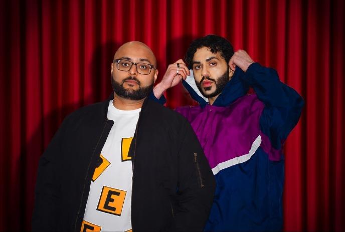 Two men stand in front of a red curtain. One wears glasses, a jacket, and a graphic tee, while the other adjusts his jacket.