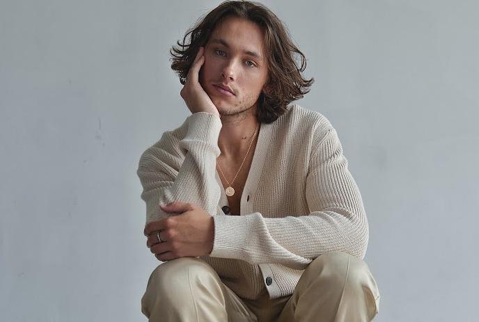 John Fellner sits thoughtfully on a stool in beige with brown shoes against a gray background.