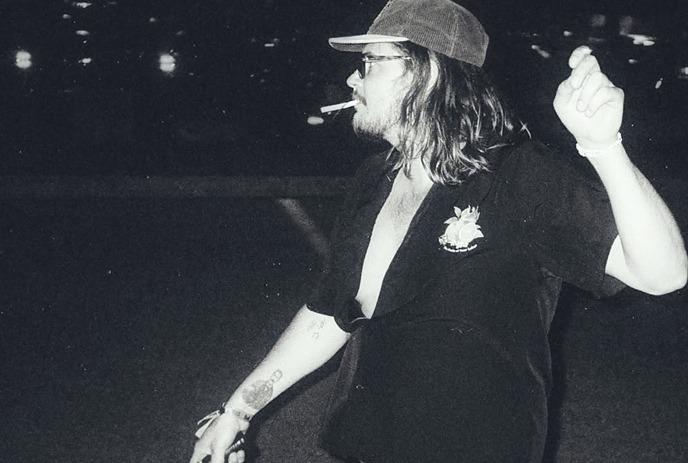 Brett Emmons with long hair and a cap swings a tennis racket energetically at night.