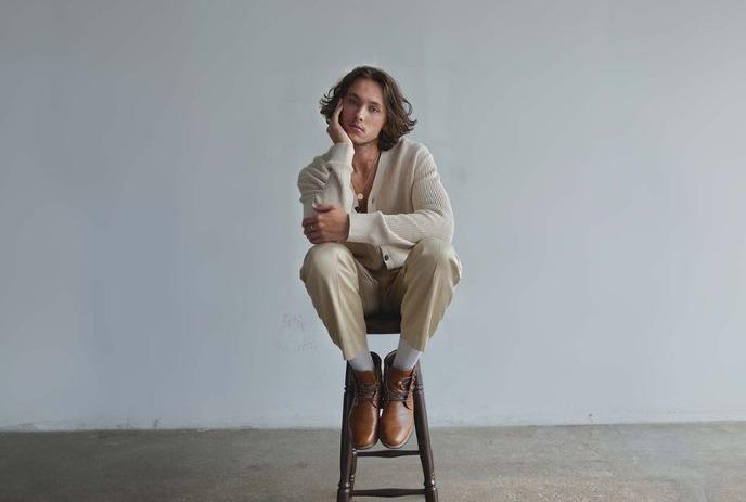 John Fellner sits thoughtfully on a stool in beige with brown shoes against a gray background.