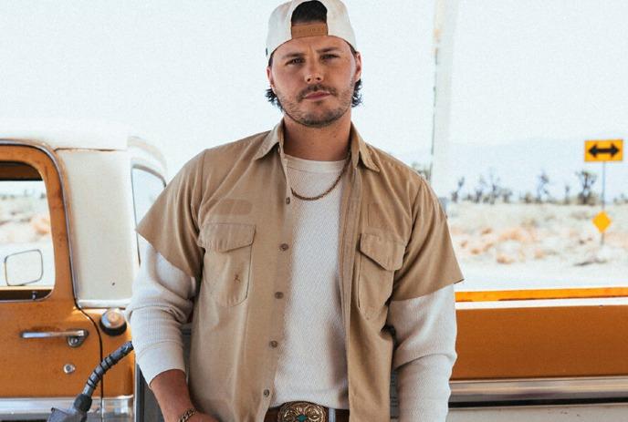 Josh Ross standing by an old orange truck in the desert, wearing a beige shirt and white cap.
