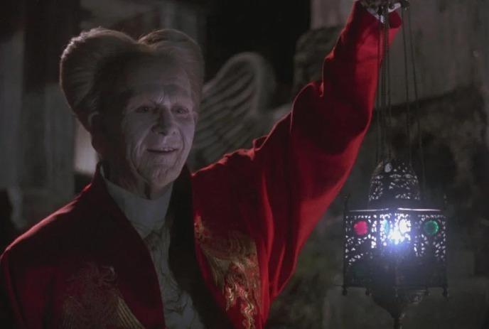 Dracula wearing a red housecoat, stands with his arm up, holding a lantern looking at something directly in front of him.
