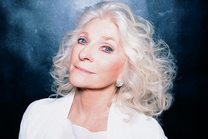 Judy Collins with expressive blue eyes and styled white hair, wearing a white outfit.