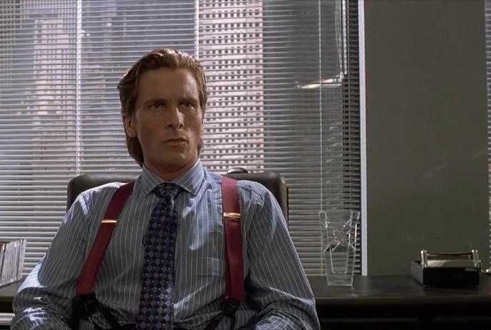 Man in office attire with red suspenders sits at desk; cityscape visible through window blinds behind.