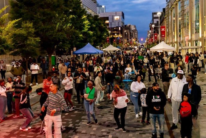 Crowd dances at Afro Fiesta Festival on lit street; tents, booths, and evening sky create festive vibe.