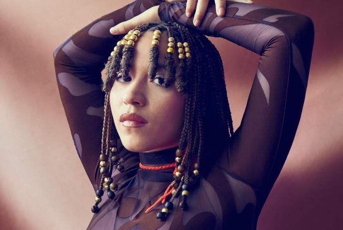 Kaia Kater poses confidently with arms raised, wearing a patterned sheer top.