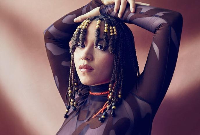 Kaia Kater poses confidently with arms raised, wearing a patterned sheer top.