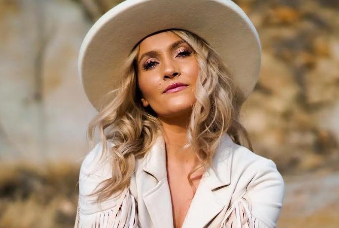 Kalsey Kulyk with long, wavy blonde hair wears a white fringed jacket and wide-brimmed hat against a rocky desert backdrop.
